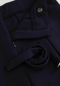 Granatowe spodnie z tkaniny o gładkiej fakturze, z podwiniętym pasem i bocznym zamkiem błyskawicznym. Zawierają czarny guzik i detal w postaci szlufki na pasek.