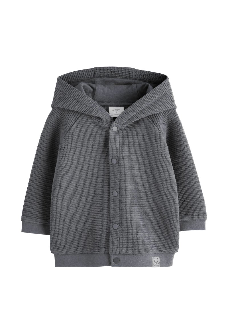 Grey waffle-textured baby hoodie with snap buttons, long sleeves, and a small fabric tag on the hem.