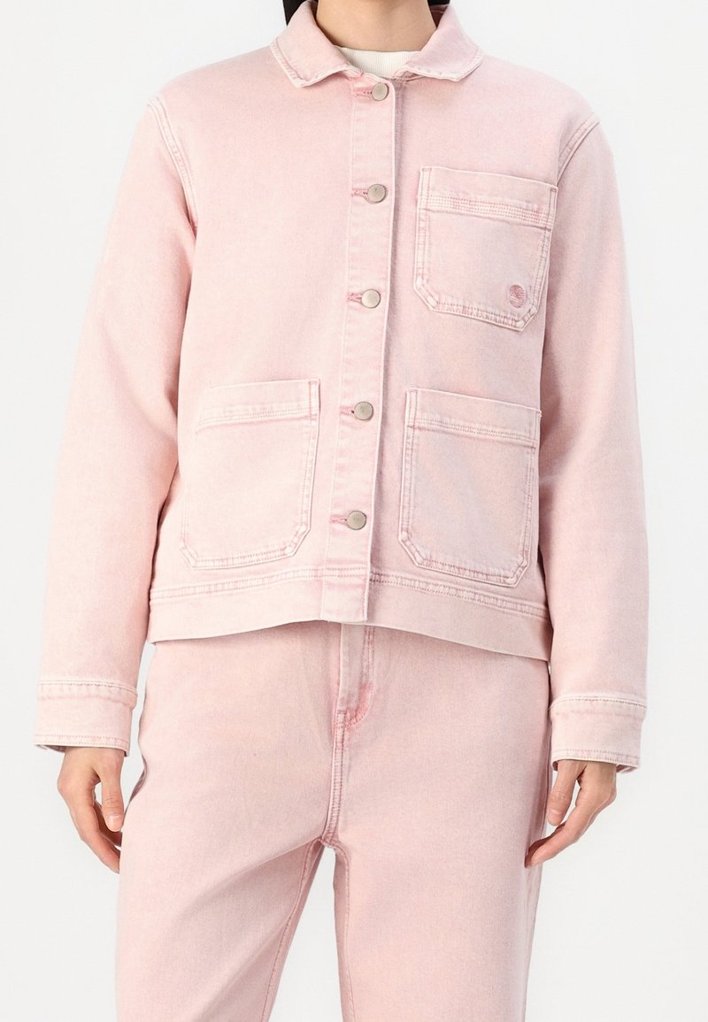 Personne portant une veste en jean rose clair avec trois poches frontales et un pantalon en jean rose assorti sur un fond blanc.
