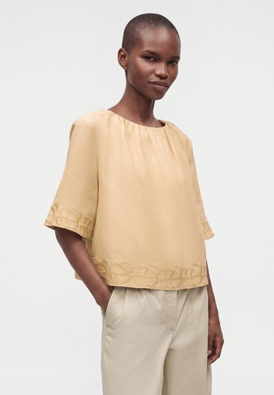 Femme portant un haut beige ample avec des boucles brodées sur les manches et l'ourlet, associé à un pantalon beige clair, debout les mains dans les poches.