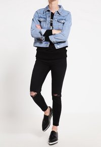 Chaqueta de mezclilla azul clara sobre un suéter negro, combinada con jeans skinny negros rasgados y zapatos negros con cordones y suelas blancas.