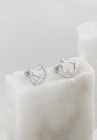 Orecchini a perno a forma di gatto argentato con design minimalista, caratterizzati da dettagli come baffi e naso. Posizionati su una piattaforma di pietra bianca texture.