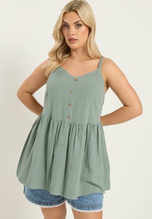 Lyshåret kvinde iført lysegrøn ærmeløs tunika med knapper over blå cowboyshorts, stående med hænderne på hoften.