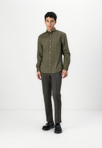 Camicia verde oliva con colletto, abbinata a pantaloni grigi a fantasia e scarpe nere. Tessuto morbido, vestibilità sartoriale, senza logo visibile.