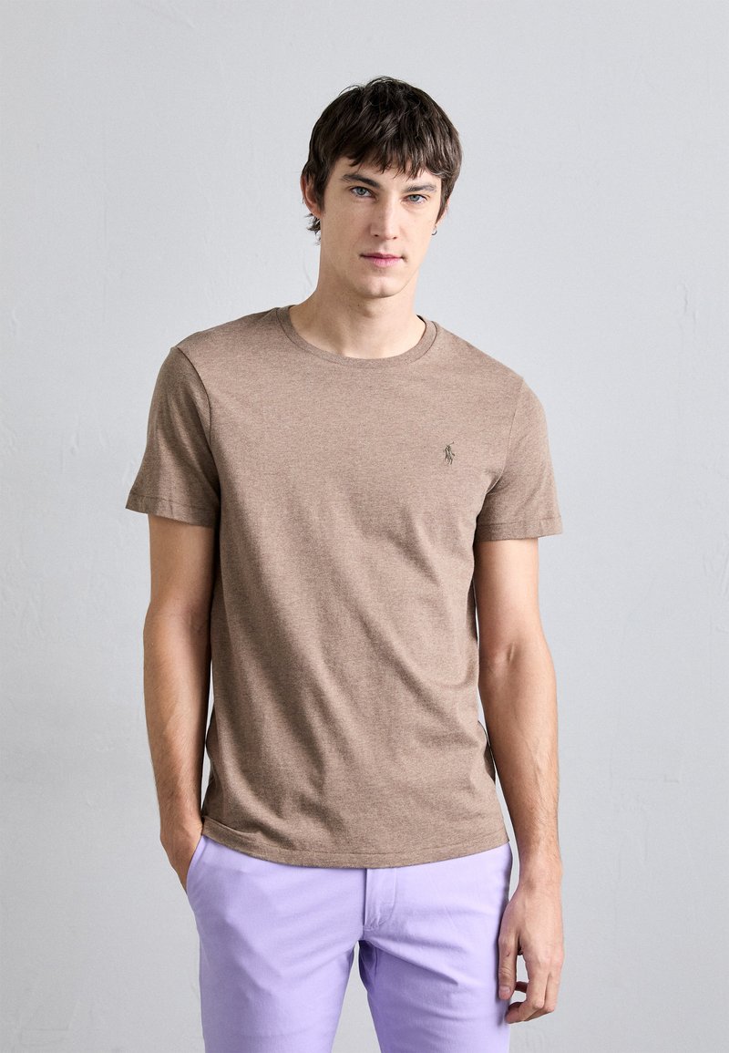 T-shirt castanho de manga curta em tecido suave e texturizado. Apresenta um decote arredondado e um pequeno logótipo no peito esquerdo, combinado com calças lilás claras.