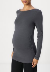 Top de maternidad gris de manga larga hecho de una tela suave, con fruncidos laterales, cuello barco y un diseño ajustado, combinado con pantalones negros de canalé.