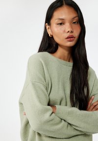 Jasnozielony, teksturowany sweter o luźnym kroju, okrągłym dekolcie i oversized rękawach. Modelka ma długie ciemne włosy i założone ręce.