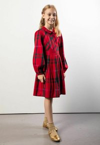 Młoda dziewczynka stoi uśmiechnięta, ubrana w czerwoną sukienkę w kratę z długimi bufkami na rękawach oraz złote buty typu Mary Jane z dopasowanymi skarpetkami.