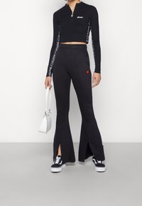 Svart kort zip-up topp med långa ärmar; matchande svarta utsvängda byxor med sprund vid anklarna; vit handväska och svarta sneakers.