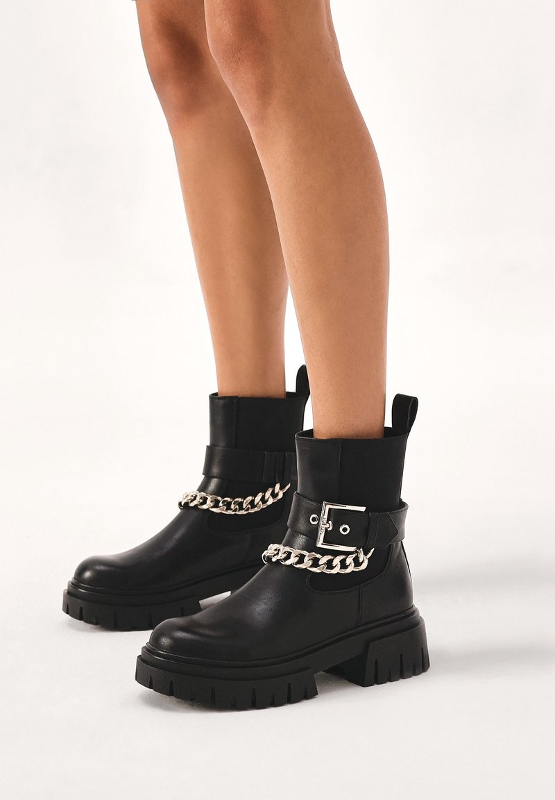Jambes portant des bottines noires épaisses avec des chaînes argentées et de grandes boucles sur un fond blanc.