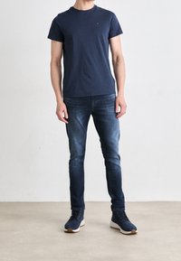 Uomo che indossa una maglietta blu navy, jeans slim-fit blu scuro e sneakers blu navy in piedi contro uno sfondo chiaro e uniforme.