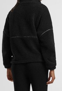 Jersey polar negro con cuello alto, presenta una superficie texturizada, hombros caídos y un detalle de costura en contraste a lo largo de la espalda.