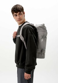 Zaino grigio su un modello, realizzato in materiale resistente con spallacci regolabili, fibbie laterali e forma cilindrica per un uso versatile.