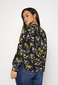 Blusa floral com mangas longas, fundo preto, flores amarelas, brancas e roxas, corte solto, design plissado nas costas. Combinada com jeans azuis.