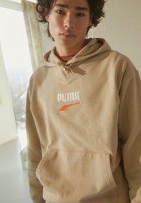 Beige hettegenser laget av myk stoff, med kengurulomme, snorhette og brodert "PUMA"-logo i hvitt og oransje.