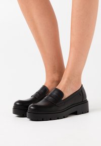 Mocassins en cuir noir à enfiler, dotés d'une semelle en caoutchouc épaisse et de détails de couture contrastés. Texture lisse, accents minimalistes.