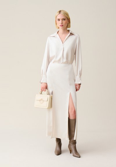 Blusa di seta beige chiaro con colletto, abbinata a una gonna lunga fino al pavimento con uno spacco alto alla coscia. Accessoriata con una piccola borsa beige.