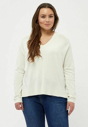 Pull à col en V couleur crème avec des manches longues, des poignets côtelés et une coupe décontractée, associé à un jean bleu foncé. Texture douce et tricotée.