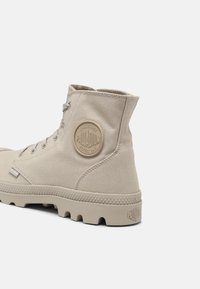 Botte montante en toile beige avec patch rond du logo Palladium sur le côté et semelle épaisse et robuste, vue de l'arrière sous un angle latéral.