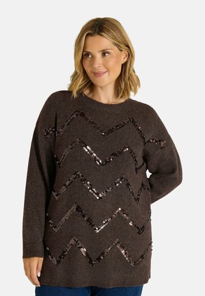 Femme portant un pull marron foncé à manches longues avec des motifs en sequins brillants en zigzag, debout avec une main dans le dos.