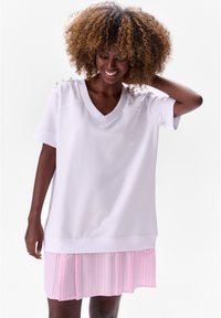 Witte V-hals tuniek met korte mouwen, voorzien van een geplooide rok met roze strepen. Inclusief zilveren schouderaccenten en een ontspannen pasvorm.