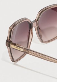 Gafas de sol Calvin Klein con montura transparente rosa, lentes cuadradas y detalles en dorado. Los lentes presentan un degradado de oscuro a claro.