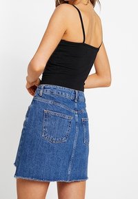 Woman wearing a black spaghetti-strap top and a blue denim skirt standing with back facing the viewer.