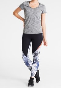 Kvinna som står med en hand på höften, iklädd en grå T-shirt med kort ärm, svarta och mönstrade leggings samt svarta träningsskor.