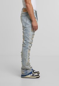 Šviesiai mėlyni nusidėvėję džinsai su įvairiais plyšimų raštais ir nusitrintais kraštais, pasižymintys siauru siluetu ir odiniu žymekliu juosmens srityje, derinami su juodais sportiniais batais.