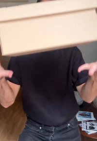 O persoană îmbrăcată cu o cămașă neagră și blugi aruncând o cutie lungă și îngustă de culoare bej în interior, cu reviste pe un masă din lemn în fundal.