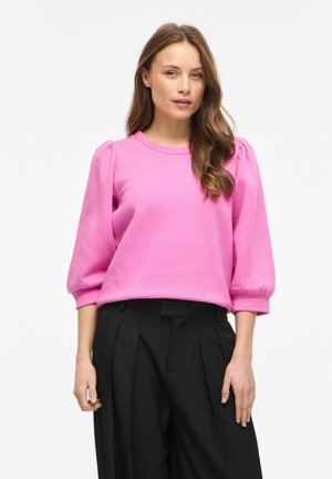 Kobieta ubrana w jaskraworóżową bluzkę z bufkami na rękawach oraz czarne plisowane spodnie z wysokim stanem, stoi na tle gładkiego białego tła.