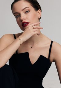 Czarna sukienka z dekoltem w serek, noszona z złotym naszyjnikiem i bransoletką z małymi czarnymi akcentami. Modelka ma włosy zaczesane do tyłu i odważną pomadkę.