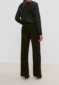 Blusa in seta nera con maniche lunghe, texture liscia, dettaglio plissettato sul retro, abbinata a pantaloni neri ampi e sartoriali, con tasche discrete.