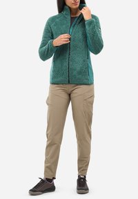 Person, der eine türkisfarbene Fleecejacke, beige Cargo-Hosen und schwarze Wanderschuhe trägt, während er die Jacke teilweise gegen einen schlichten weißen Hintergrund zippt.