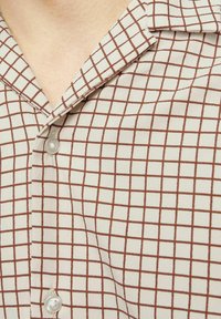 Chemise à manches courtes avec boutonnière en tissu léger, présentant un fond beige avec un motif à carreaux marron et un col en V.