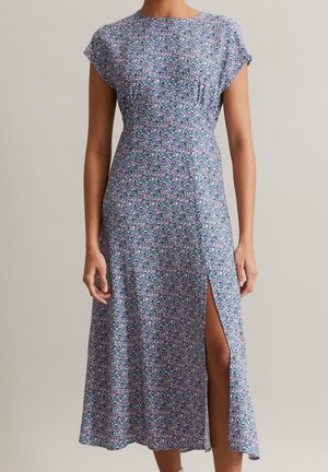 Mujer vistiendo un vestido midi morado con flores, mangas cortas y una abertura alta en el muslo sobre un fondo neutro.