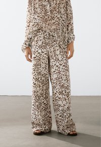 Lehký béžový set z látky, který obsahuje volně střiženou košili s dlouhými rukávy a široké kalhoty, oboje s hnědým tečkovaným vzorem.