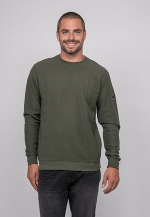 Homme souriant, portant un pull à manches longues vert olive et un pantalon foncé, debout devant un fond uni gris clair.