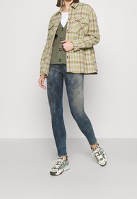 Grön och gräddfärgad rutad oversized skjortjacka, lager på lager över en grön knäpptröja och vit t-shirt, ihop med blå tajta jeans och mönstrade sneakers.