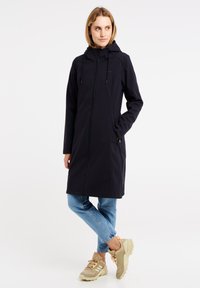 Lungo cappotto nero con cappuccio, dotato di zip frontale e tasche laterali, abbinato a jeans azzurri e sneakers beige con dettagli testurizzati.