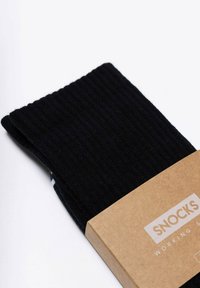 Czarne prążkowane skarpetki zapakowane w etykietę z brązowego kartonu, na której widnieje napis "SNOCKS" w białym kolorze, podkreślając prosty i funkcjonalny design.