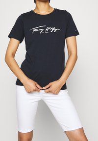 Námořnická modrá bavlněná tričko s krátkým rukávem, zdobené stříbrným potiskem loga, které zní "Tommy Hilfiger" a rok "1985." V kombinaci s bílými šortkami.