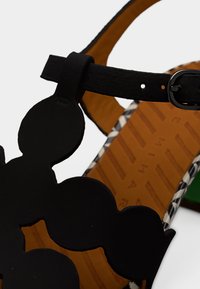 Μαύρο σανδάλι με κυματιστές εγκοπές, χαρακτηριστικό του ένα υφασμένο λουρί και αγκράφα. Η σόλα είναι καφέ με κάθετες αυλακώσεις και πράσινη λεπτομέρεια.