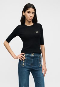 Haut noir côtelé à manches courtes avec une petite poche et un accent métallique. Porté avec un pantalon en denim bleu orné de boutons dorés sur le côté.