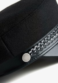 Svart texturerad keps med ett flätat läderband och en rund silverknapp på sidan, visas i nära detalj.