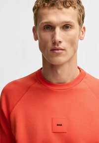 Roter Pullover mit geripptem Kragen, Raglanärmeln und einem quadratischen Logo-Patch in dunklerem Rot mit eingeprägtem Text. Glatte Textur, lässiges Design.