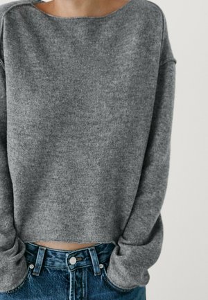 Person wearing a loose gray knit sweater and blue jeans with hands tucked into pockets against a plain background.