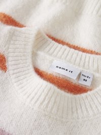 Pleteni pulover z okrolim izrezom, ki je narejen iz mehkega belega materiala z oranžnim črtastim vzorcem. Oznake prikazujejo velikost in blagovno znamko.