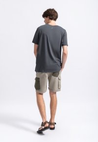 Grijze katoenen T-shirt, relaxed fit. Olijfgroene cargoshorts met zijzakken. Zwarte sandalen met een band. Neutrale achtergrond.