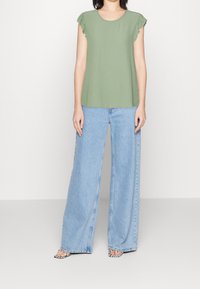 Zielony krótki top z falbaniastymi rękawami, w zestawie z szerokimi niebieskimi dżinsami i wzorzystymi sandałami. Prosty, luźny krój i design.